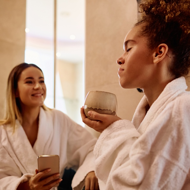 Zwei Personen sitzen in Bademänteln in einem Spa‑Bereich und halten Getränke in den Händen.