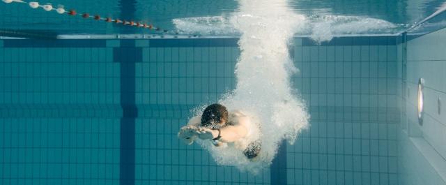 Eine Person taucht in ein Schwimmbecken ein, unter Wasser sind Luftblasen und die ausgestreckten Arme sichtbar.