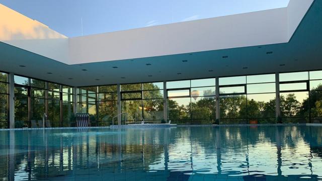 Blick über ein Schwimmbecken in einer Halle mit großen Fensterflächen und Ausblick ins Freie und offenem Dach.