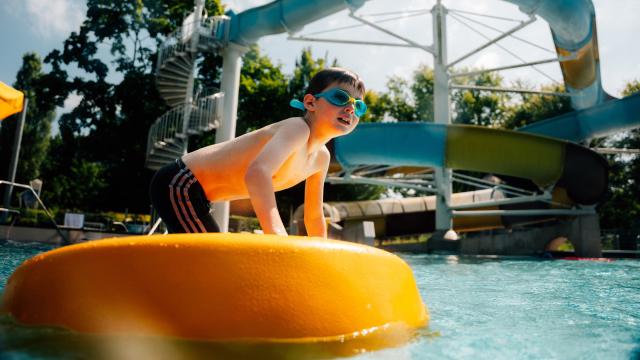 n Kind mit Schwimmbrille stützt sich in einem Freibadbecken auf einem gelben Schwimmelement ab, im Hintergrund ist eine Wasserrutsche zu sehen.