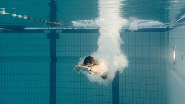 Eine Person taucht in ein Schwimmbecken ein, unter Wasser sind Luftblasen und die ausgestreckten Arme sichtbar.